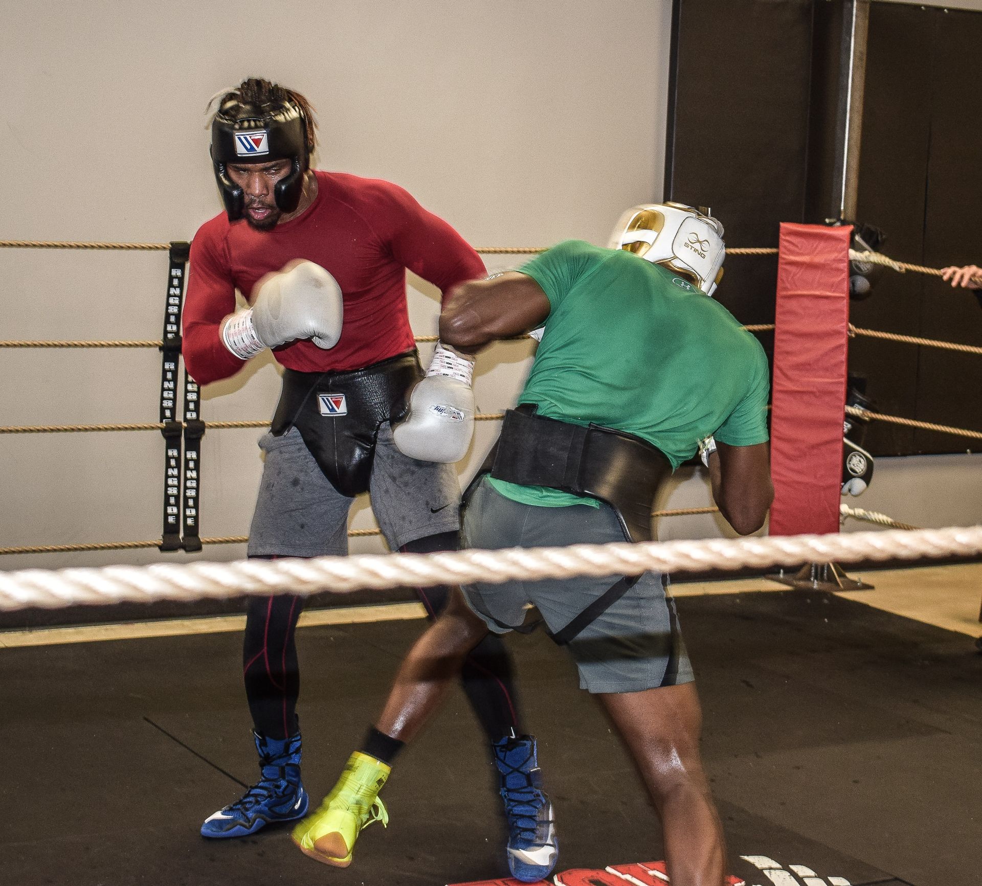 Boxers sparring in a ring, one in red throwing a punch at the boxer in green. They wear headgear and protective gear.