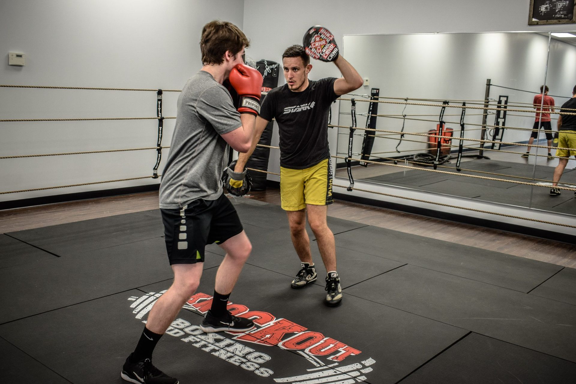 Two men boxing in a gym, one wearing red gloves and one with mitts, standing on a boxing mat.