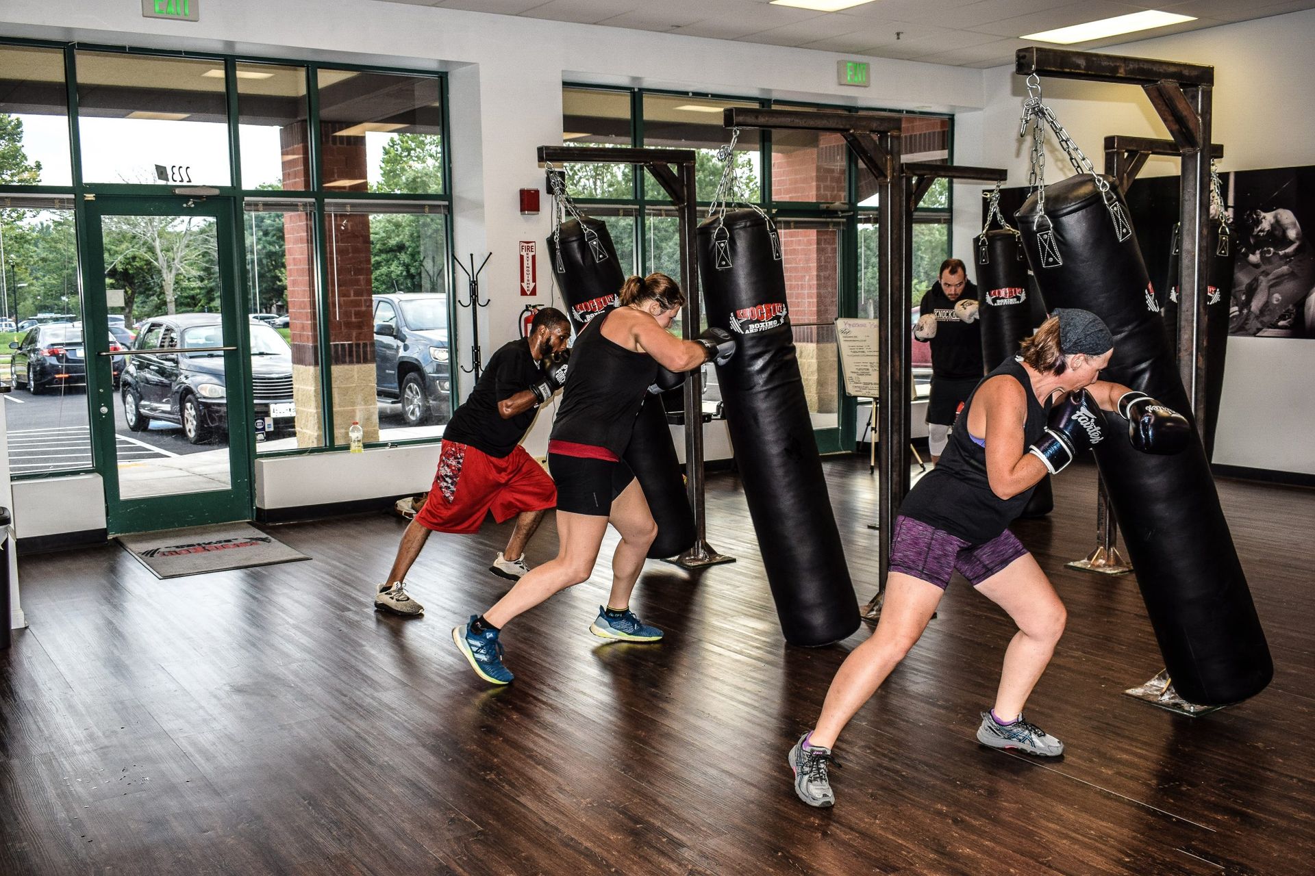 People in a boxing gym, working out with punching bags. Dark wood floor and windows.