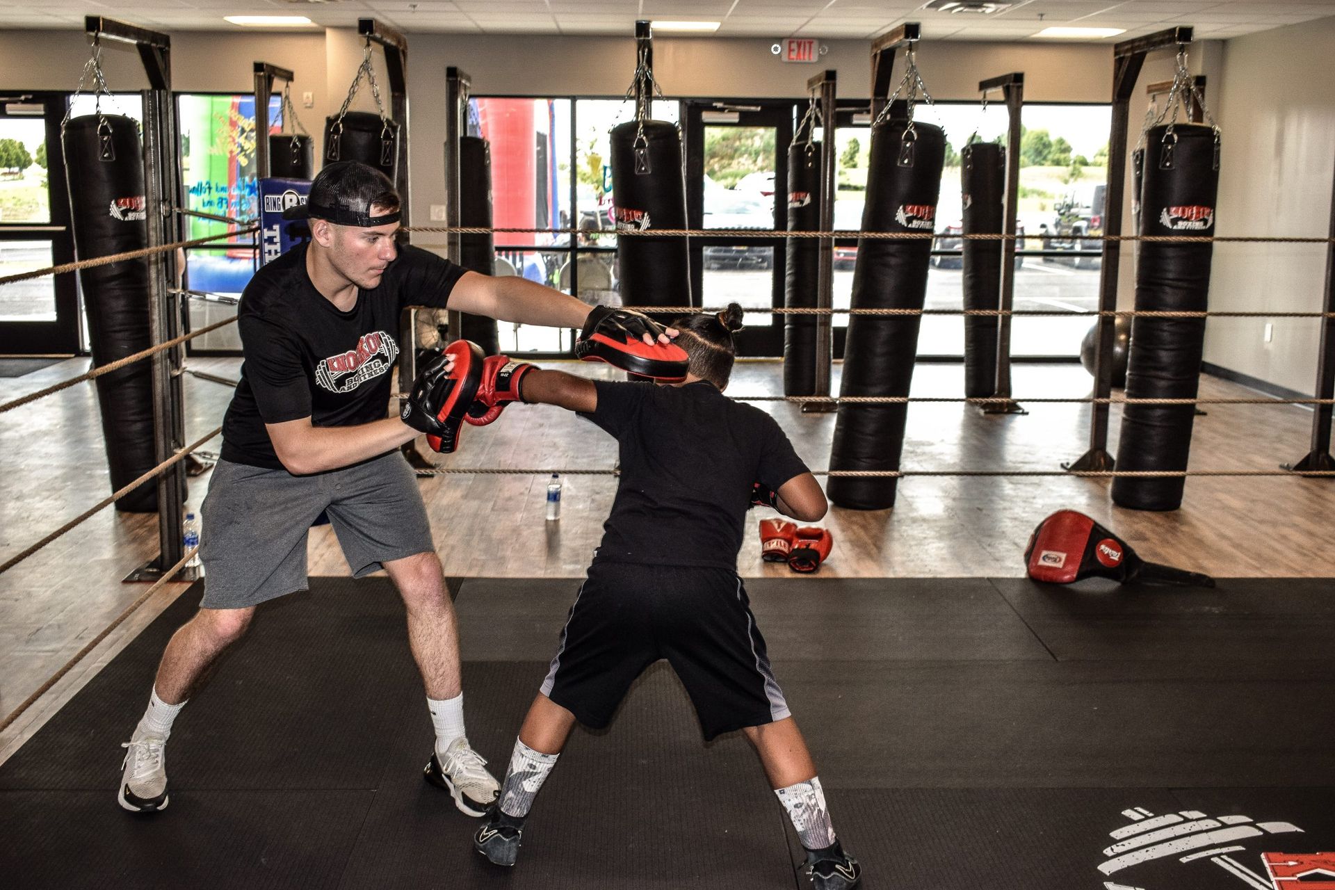 Man and boy boxing in a ring; man in black shirt, boy in black shorts, gym setting.