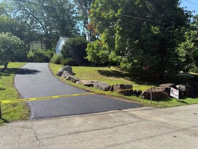 Newly paved asphalt driveway with a yellow caution tape barrier, flanked by large landscaping rocks and trees.