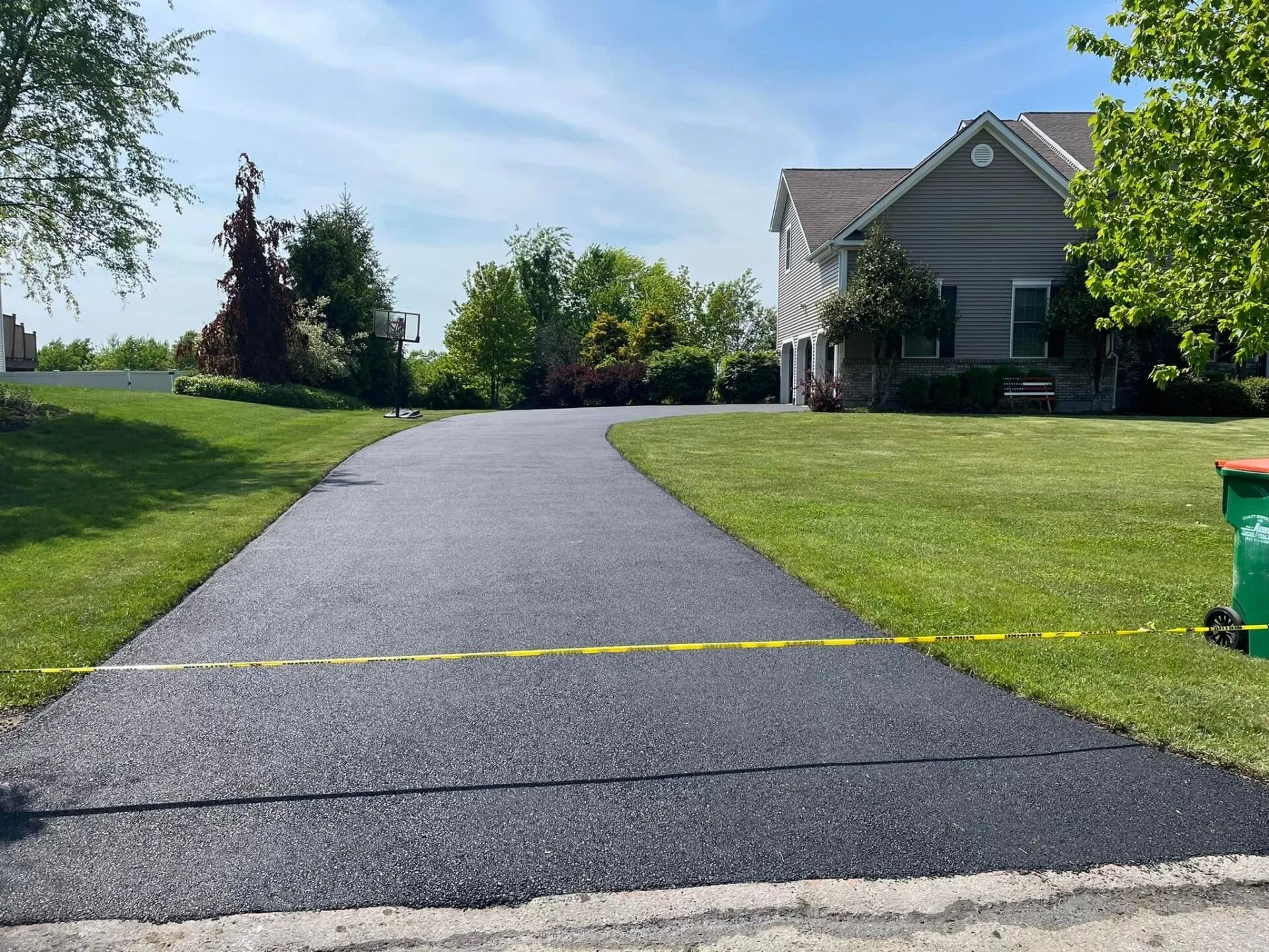 A freshly paved asphalt driveway leads to a suburban house on a sunny day, with yellow caution tape across the entrance.