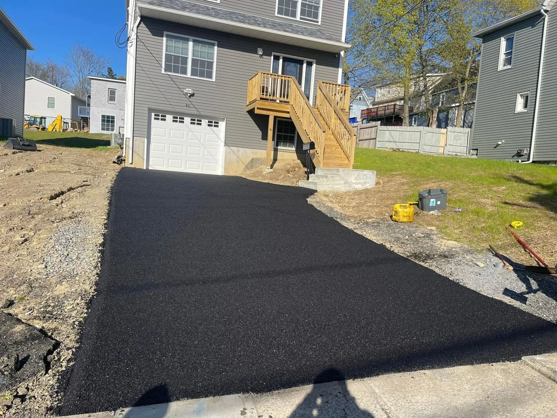 A newly paved asphalt driveway leads to the garage and stairs of a gray residential house on a sunny day.