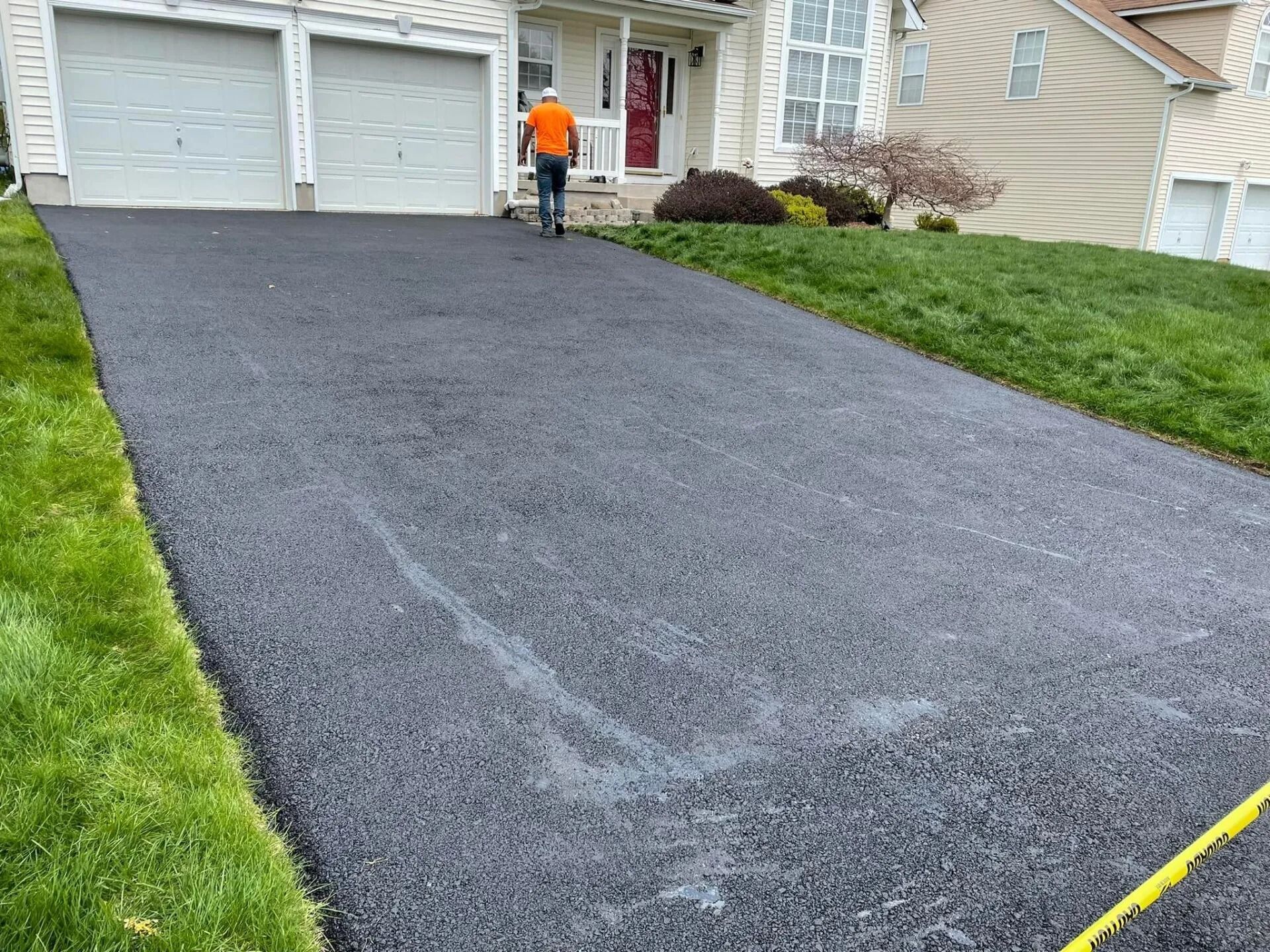 A worker in an orange shirt stands on a freshly paved dark asphalt driveway in front of a suburban house.