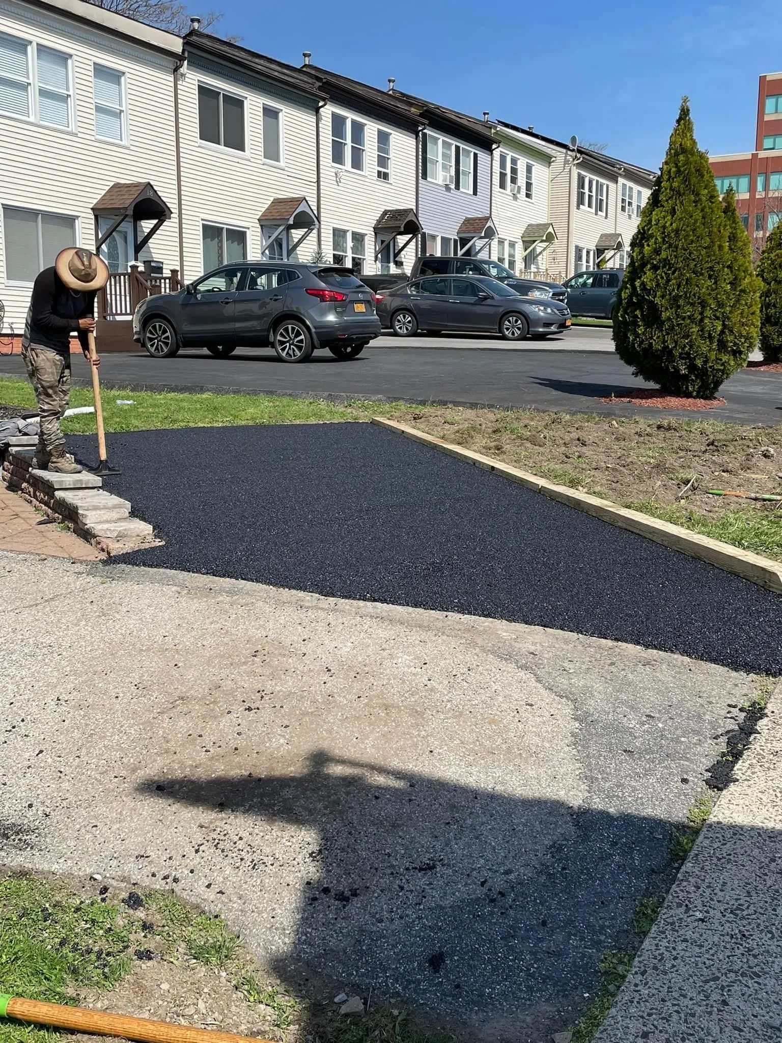 A person spreads dark mulch in a garden bed in front of a townhouse complex with a parked car nearby.
