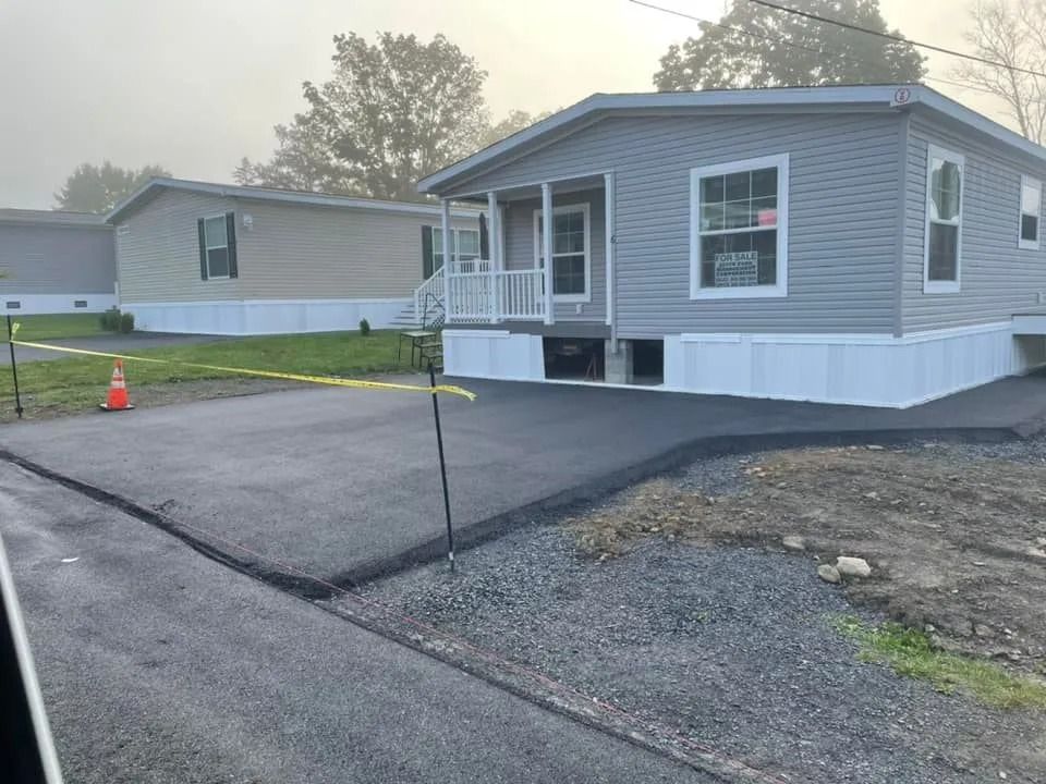A gray mobile home with a newly paved asphalt driveway and a yellow caution tape stretched across the front.