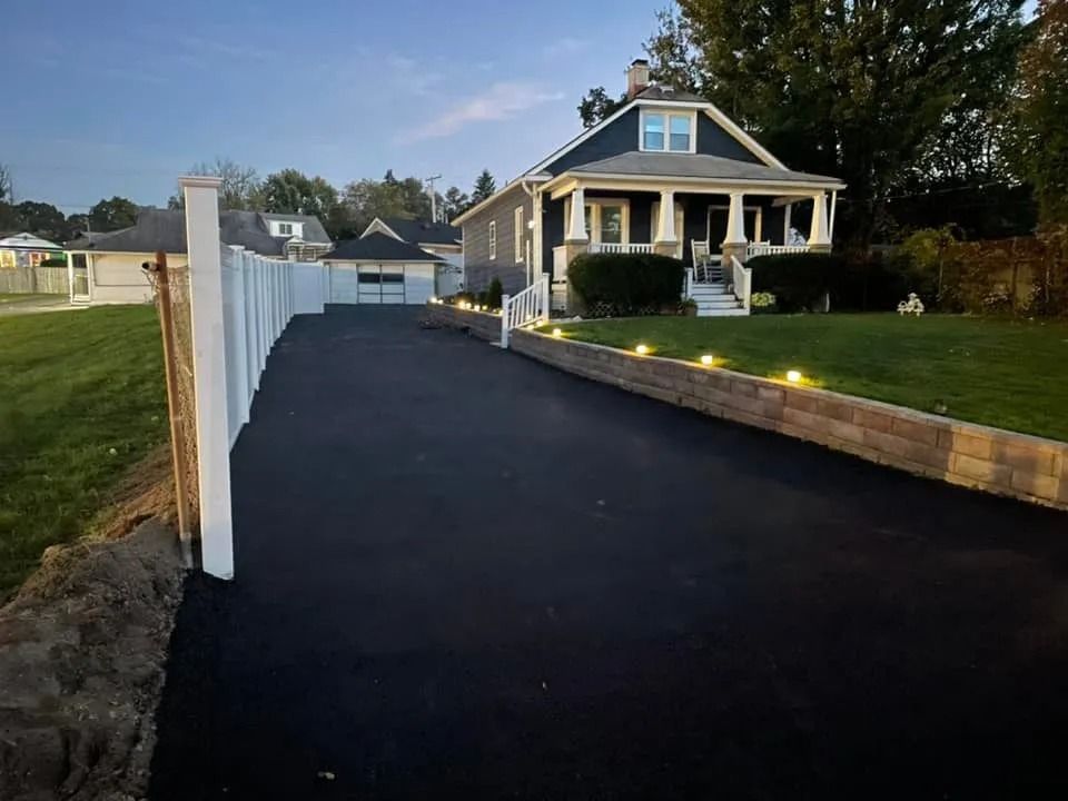 A freshly paved black asphalt driveway leads to a gray house with a covered porch, bordered by a white fence and lights.