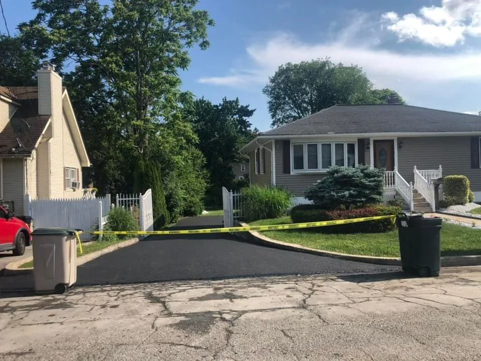 A fresh asphalt driveway between two houses, blocked off by yellow caution tape with trash bins on either side.