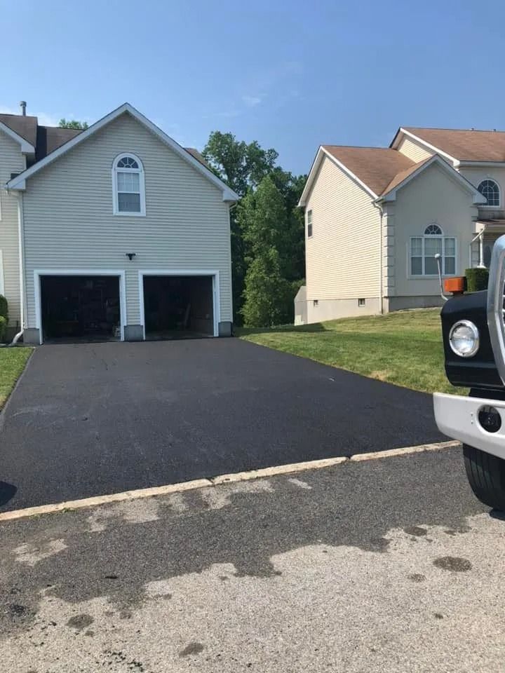 A freshly paved black asphalt driveway leads to a two-car garage of a light-colored house on a sunny day.