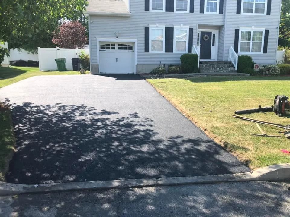 A newly paved asphalt driveway leads to the garage of a light gray two-story suburban house on a sunny day.