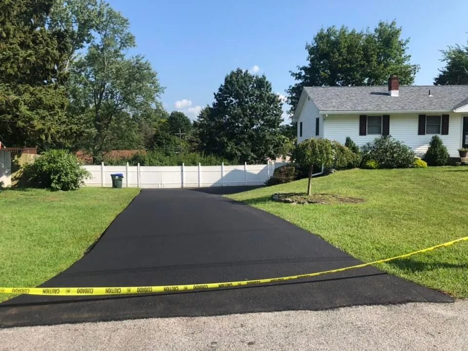 A freshly paved, dark asphalt driveway leads toward a white single-story house behind a white fence under a clear blue sky.