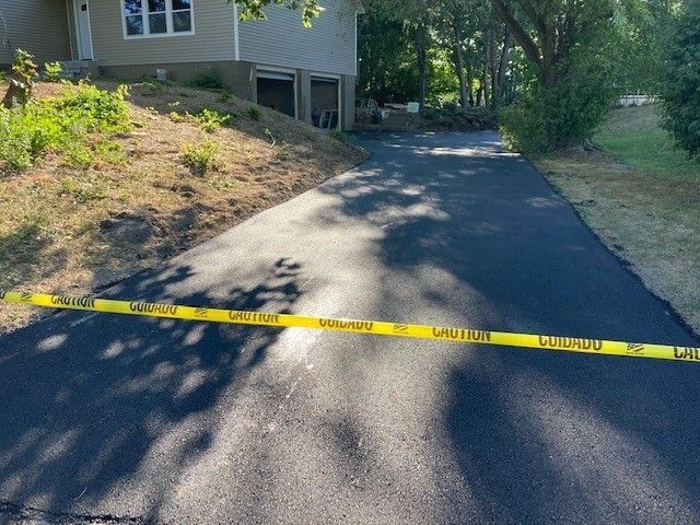 A freshly paved dark asphalt driveway leads to a suburban home, blocked off by yellow caution tape.