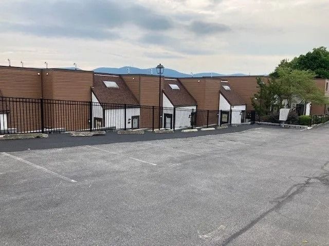 An asphalt parking lot in front of a row of modern, brown, multi-level townhomes under a cloudy sky.