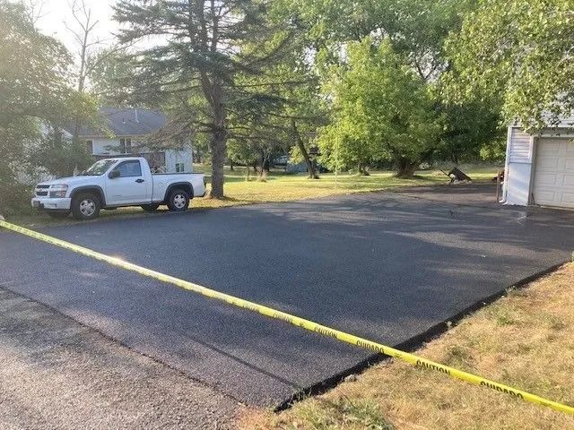 A freshly paved asphalt driveway sits in front of a house, blocked off by yellow caution tape with a white truck parked.