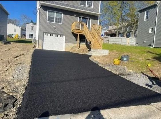 A freshly paved dark asphalt driveway leads to the garage and front wooden stairs of a grey, multi-story suburban house.