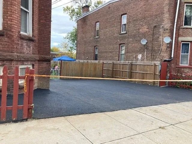 Freshly paved black asphalt driveway between two brick apartment buildings, blocked off by a red fence and yellow tape.