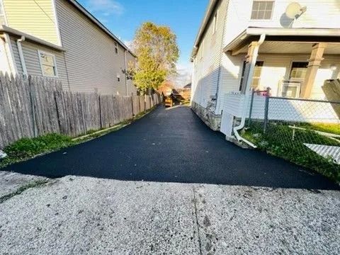 A freshly paved asphalt driveway sits between two houses, extending from a concrete foreground toward a distant tree.