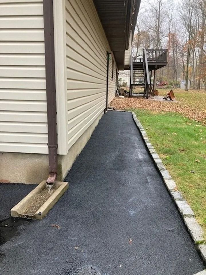 An asphalt path runs along the side of a beige house, featuring a downspout drainage stone and a stone border.