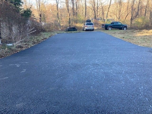 A newly paved black asphalt driveway leading toward two parked vehicles in a wooded area.