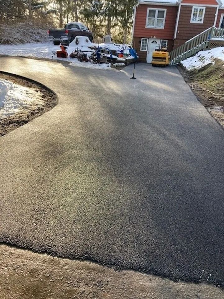 Newly paved asphalt driveway leading to a red house with snowy surroundings and construction equipment in the background.