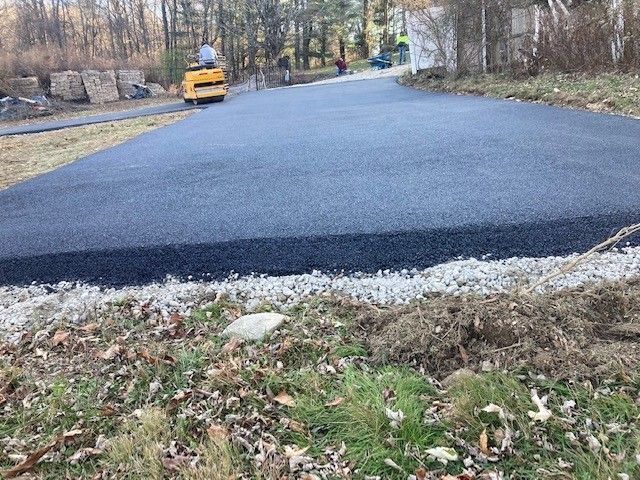 A freshly paved dark asphalt driveway with a yellow construction roller in the background on a gravel base.