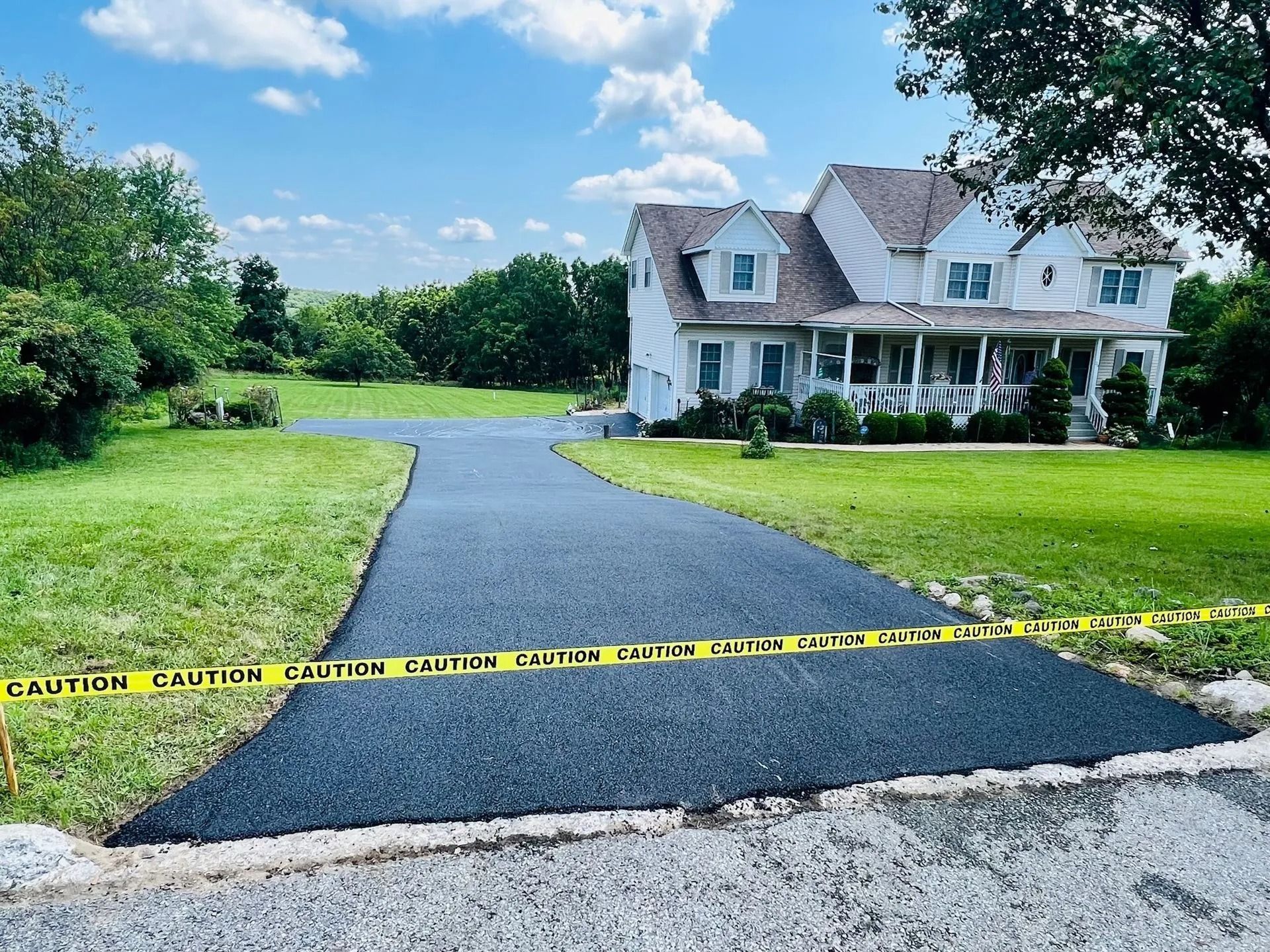 A newly paved black asphalt driveway leads to a white house with a covered porch, blocked by yellow caution tape.