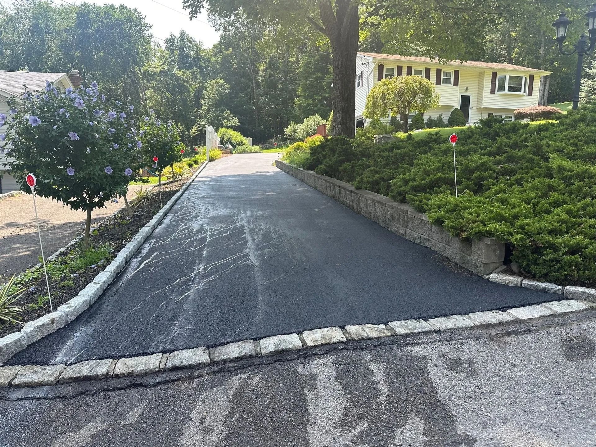 A newly paved asphalt driveway with stone-block edging leads up a hill to a house surrounded by trees and shrubs.
