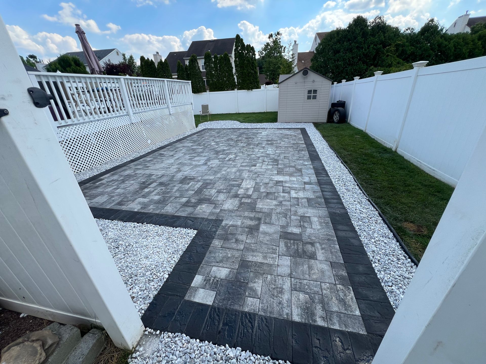 A white fence surrounds a brick driveway in a backyard.