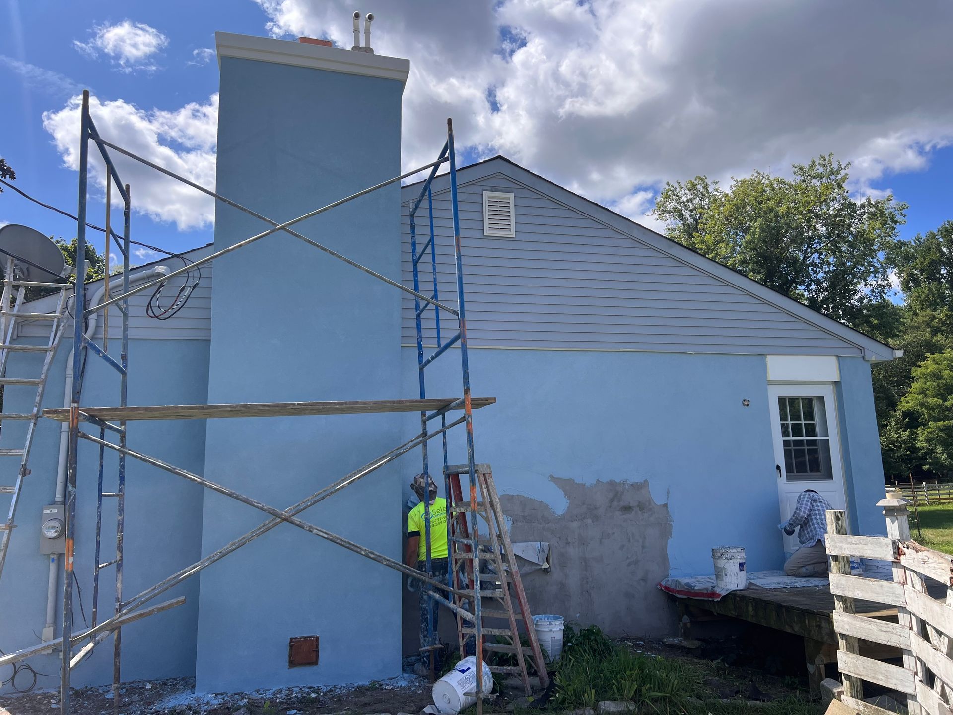 A man is standing on a ladder in front of a blue house being painted.