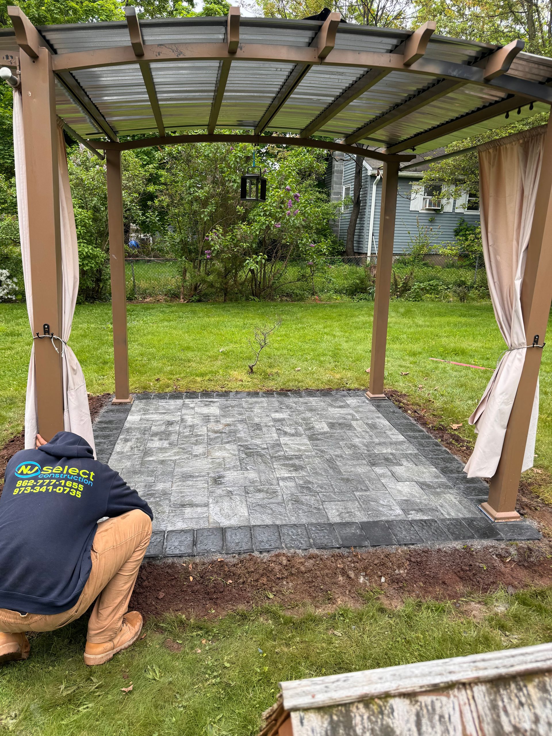 A man is kneeling under a pergola in a backyard.