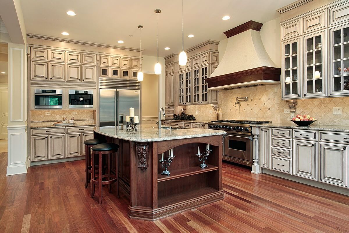 Luxury kitchen featuring a granite island, custom cream cabinets, a dark wood hood, stainless steel appliances, and wood.