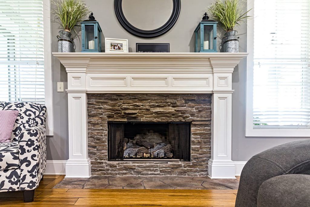 A white fireplace mantle with a stone surround, topped with a round mirror, lanterns, and greenery in a living room.