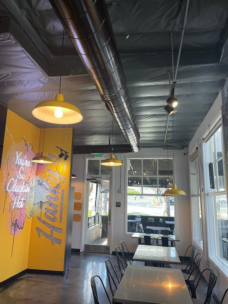 Interior view of a cafe with yellow accents, tables, and pendant lights. A mural and windows are also visible.