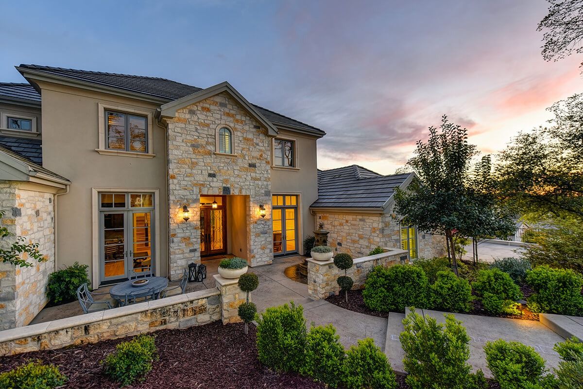 Luxury home exterior with stone facade, manicured landscaping, and sunset sky.