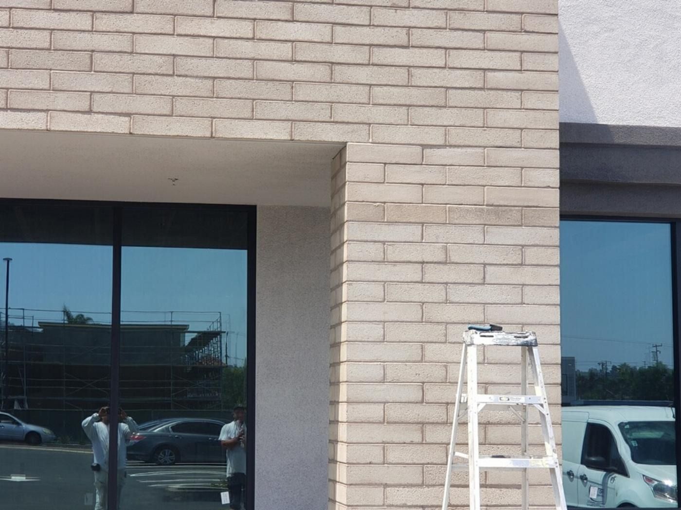 Exterior view of a building with brick facade and glass windows reflecting the outside. A ladder stands near the wall.