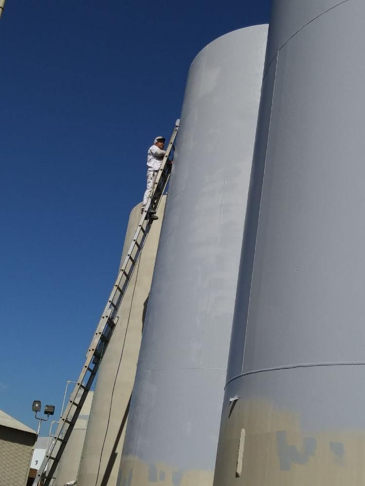 Person on a ladder painting a tall cylindrical industrial tank gray against a blue sky.