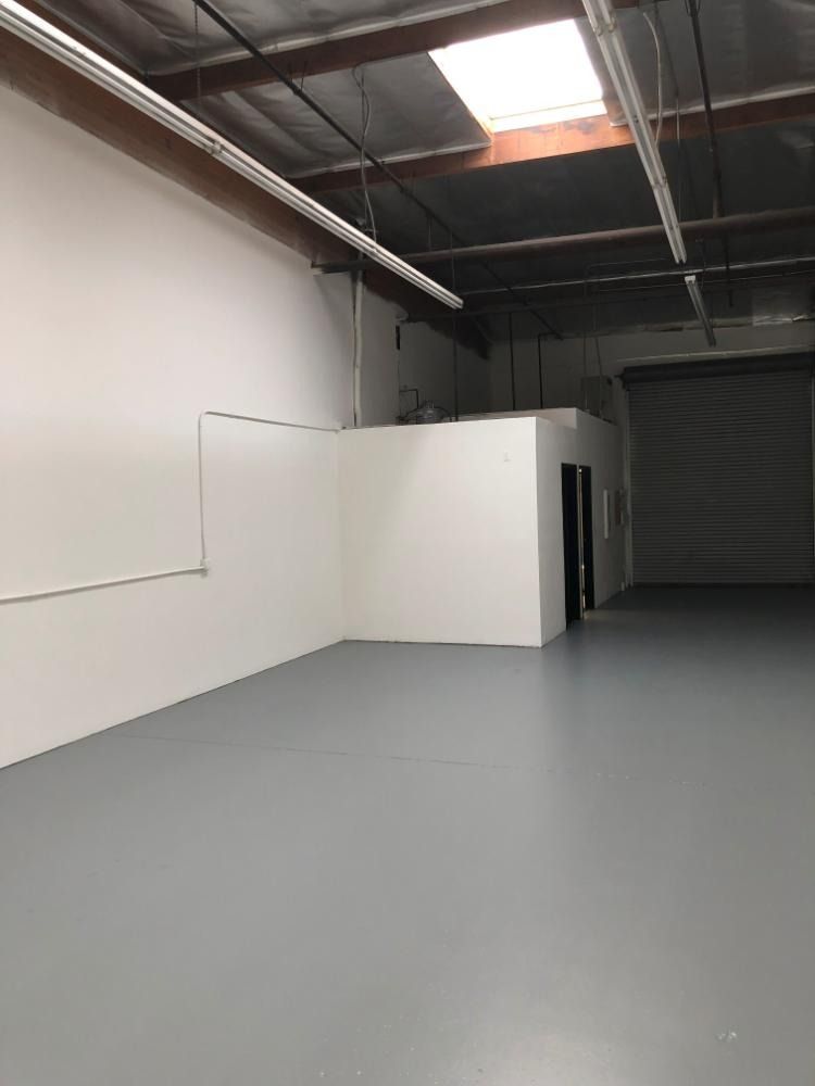 Empty warehouse interior with white walls, gray floor, overhead lighting, and a small office space.
