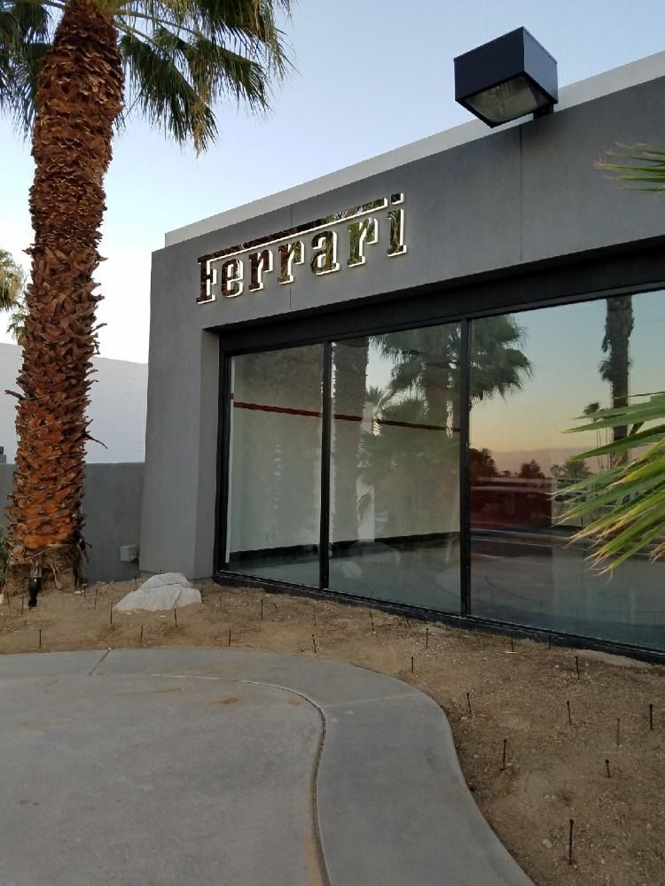 Ferrari dealership storefront with large windows, gray facade, and palm trees.
