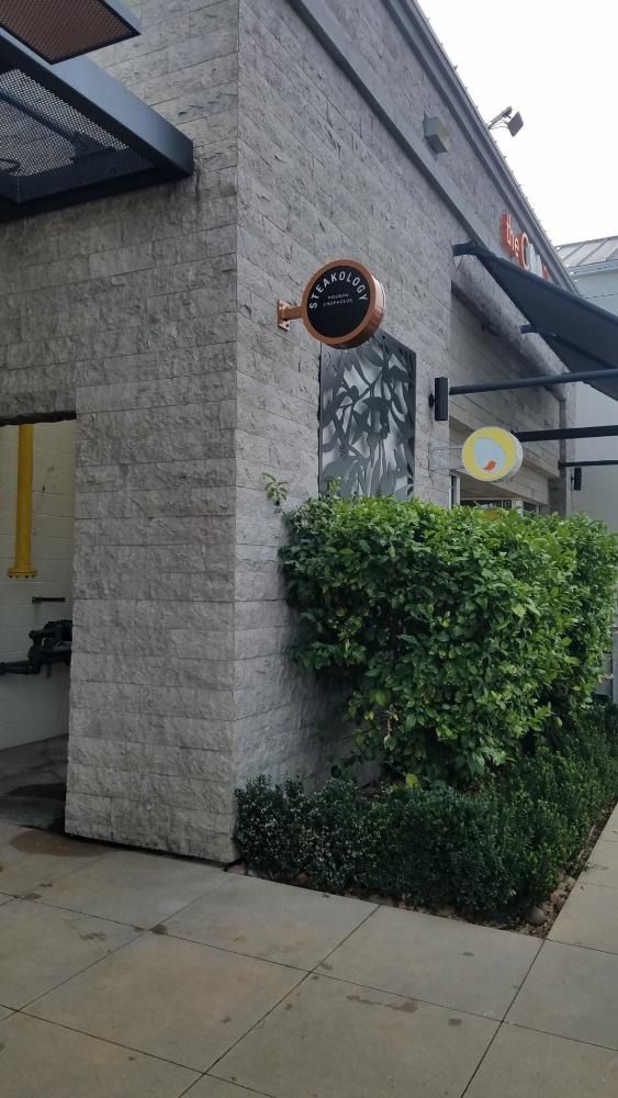 Exterior of a restaurant with a brick facade and round sign; a green bush sits below.