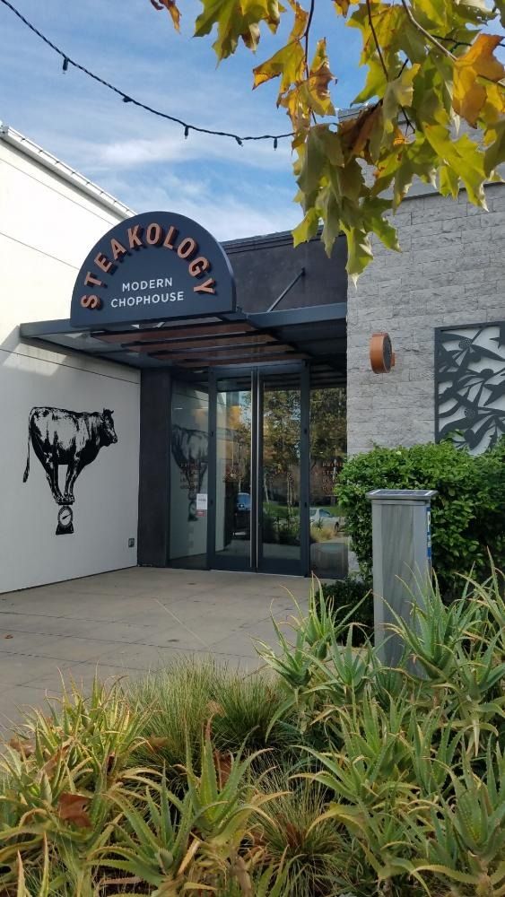 Steakology restaurant entrance with sign, glass doors, and cow mural. Green foliage and blue sky are visible.