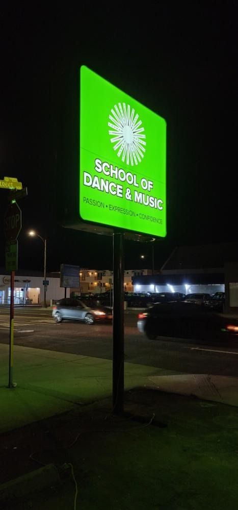 Green sign at night: School of Dance & Music with white text, starburst logo. Cars in background.