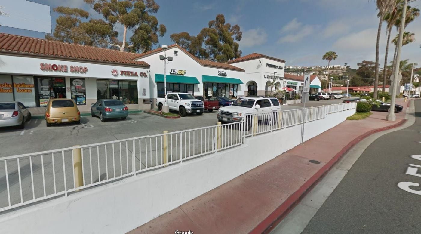 Shops in a strip mall with a white wall and street. Sunny day with cars parked.