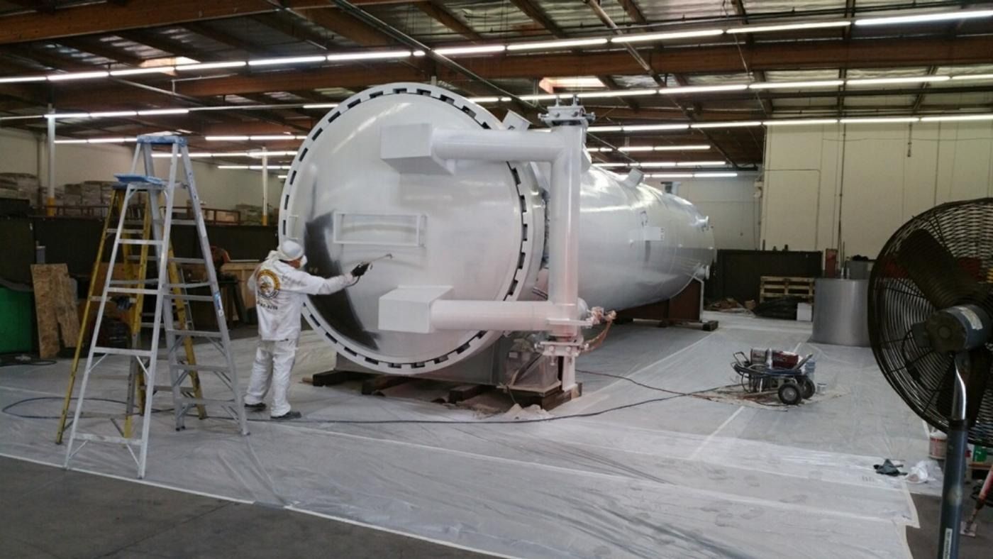 Person in protective suit painting a large white industrial tank indoors.