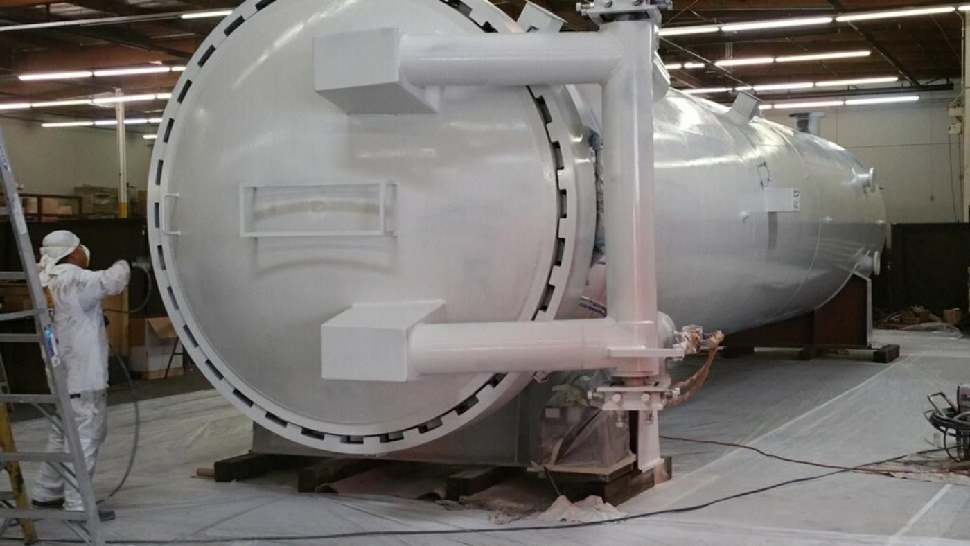 Person in white suit painting a large white industrial pressure vessel.