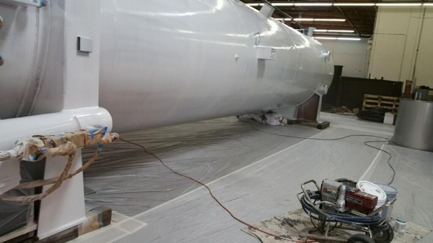 Large, white industrial structure being painted in a factory. Paint sprayer is in the foreground.