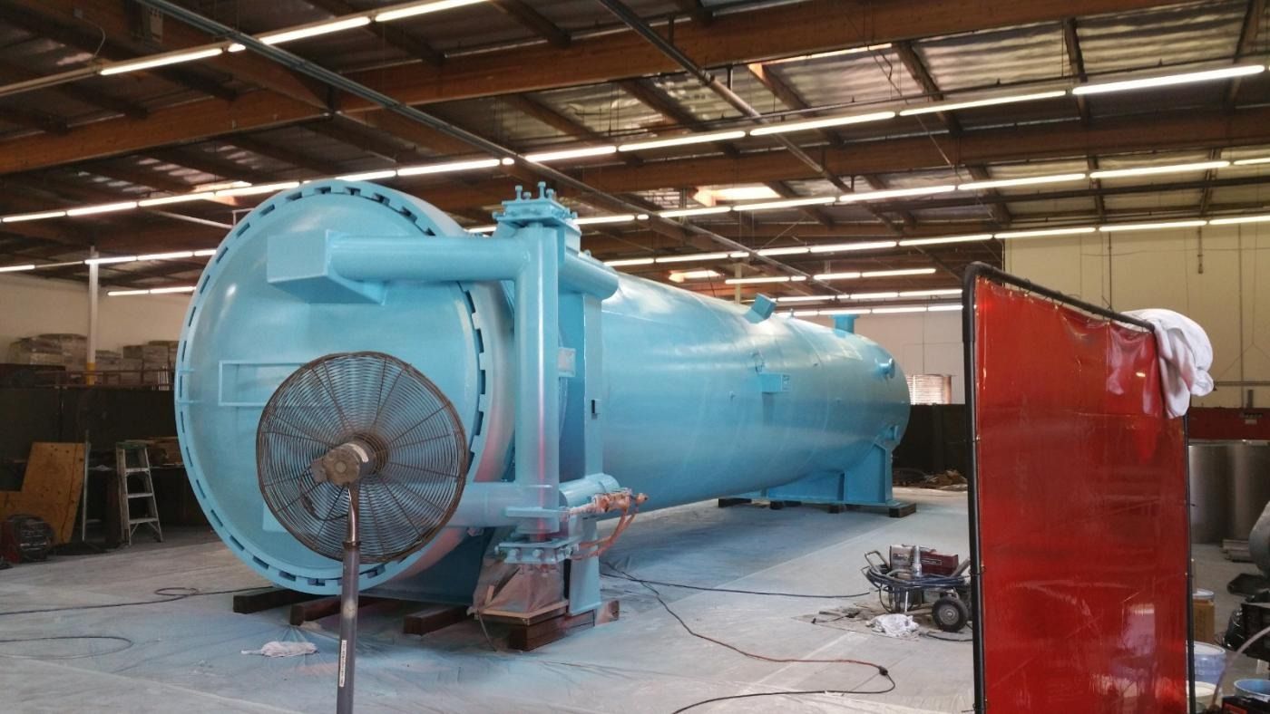 Large, light blue industrial tank with attached pipes, inside a workshop. A large fan is in front.