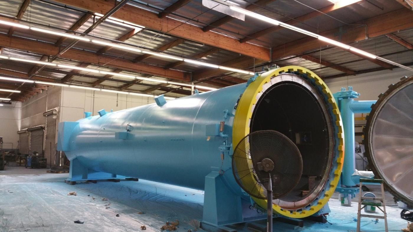 Large, blue industrial autoclave with open end, yellow trim, inside a warehouse.