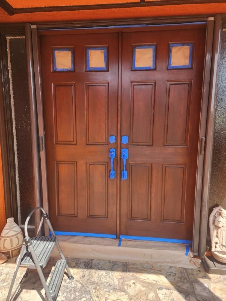 Double wooden front doors, stained brown, with blue painter's tape and handles, and a small ladder.