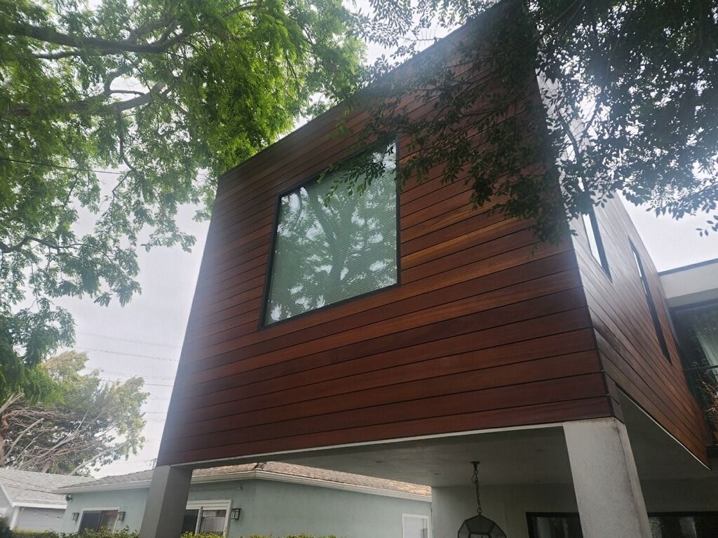 Modern house exterior with brown wood siding, large window, and partial view of trees.