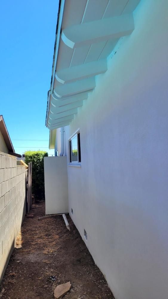 White building with gutters, a small window, and a narrow dirt path next to a brick wall.